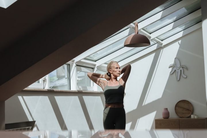 A woman standing in a sunlit attic, arms behind her head, exuding tranquility.