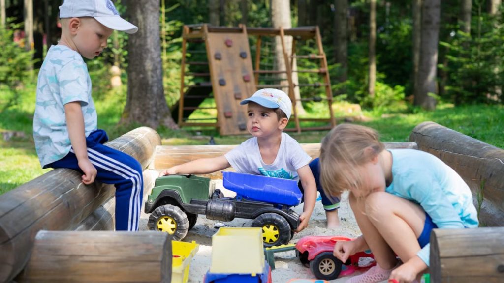 Three children engaged in imaginative play in an outdoor sandbox with toys.