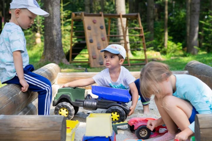 Three children engaged in imaginative play in an outdoor sandbox with toys.