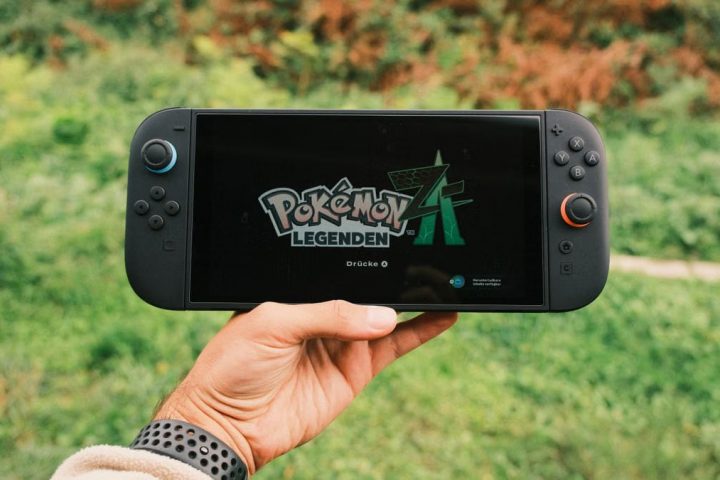 A hand holding a handheld gaming console displaying the Pokémon Legends game screen outdoors.